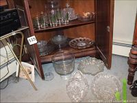 Wide shot showing various crystal and pressed glassware items inside and outside of a wooden cabinet, including plates, bowls, and a cake plate.