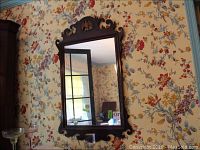 Full view of rectangular wood framed wall mirror with ornate carved top against floral wallpaper