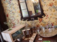 Overall display of the barware including decanters, iridescent stemware, margarita glasses, ceramic wine glasses, and porcelain bottle name chains on a wooden table with floral wallpaper background.