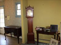 Full view of tall vintage Smiths hall clock standing between two wooden furniture pieces in a room with yellow walls and windows.