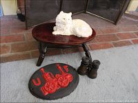 Photo showing white cast iron cat doorstop on small wooden stool, vintage binoculars and cast iron fire pumper plaque on floor in front of fireplace.