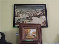 Two landscape artworks on wall: smaller framed original autumn oil painting in rustic frame positioned below larger winter landscape print in black frame.
