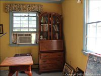 Full view of the vintage wooden secretary desk placed against a yellow wall between two windows, showing two glass doors on top and the overall height and shape.