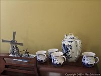 Full view of four Delft mugs and the Delft ceramic teapot on a wooden surface against a yellow wall.