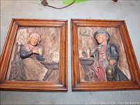 Photo showing the pair of plaster bas relief plaques featuring a man and a woman seated at tavern tables, framed in wood with visible paint wear.