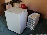 Side view of off-white metal two-drawer filing cabinet and several white letter trays stacked beside it on carpeted floor.