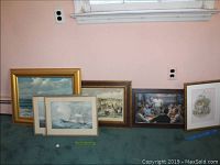 Six pieces of framed wall art leaning against a wall on carpeted floor including maritime scenes, group portraits, and a child with faeries.
