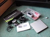 Photo showing Bose Soundlink speaker with manual, various cables, flip phones and accessories in a plastic bag, and Apple external drive on green carpet.