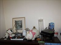 Surface displaying many assorted items including figurines, piggy banks, a glass lamp base, framed print, jewelry trays and others