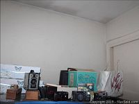 Photo showing various vintage cameras including box-style and compact cameras along with storage boxes and other items on shelves against a wall.