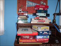 Stack of vintage board games on a small desk against an orange wall, including Parcheesi, Monopoly, Duell, Tri-Ominos, Trivial Pursuit, Pictionary, Olympic Soccer Game, and others.