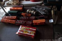 Top-down view of multiple vintage model train cars in orange, green, maroon, and white colors, along with metal train tracks and the boxed donkey cigarette dispenser on a table.