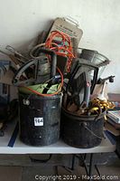Two black buckets filled with assorted used yard tools including electric hedge trimmers and hand sprayers.
