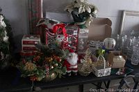 Holiday decorations on a table including wrapped boxes of ornaments, Santa figurines, two baskets, a Christmas wreath with pine cones and berries, a small faux flower pot with white poinsettias, and a gold wire Christmas tree figure.