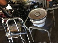 Photo showing silver walker, portable commode with white bucket and lid on gray chair, black mobility scooter partly visible behind them.