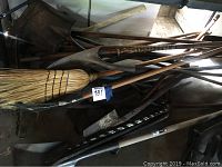 Photo showing a collection of yard tools including brooms, shovels, and pitchforks piled together, wooden handles and metal heads visible, some rust and wear present.