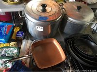 View of large aluminum pots with lids, square copper frying pan and other assorted cookware on a table showing wear.