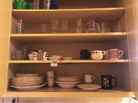 Wooden shelves holding Mikasa plates and bowls, clear glassware, coffee mugs, salt and pepper shakers arranged across three shelves.
