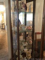 Glass cabinet with multiple shelves showing ceramic teacups, saucers, decorative boxes, and figurines inside.