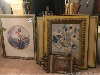 Five framed pieces stacked against a wall showing floral and landscape art.