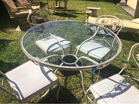 Round outdoor metal table with glass top shown with 4 matching metal chairs on grass outdoors.
