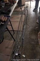 Photo shows six long heavy duty metal chains laid on floor and a cardboard surface, each chain has thick metal links and a hook on the end.