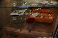 Assortment of formal servingware displayed inside a glass cabinet including glass punch bowl, cake plates, decanter, and boxed demitasse coffee cup set.