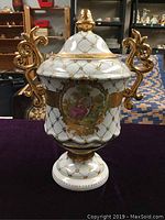 Full frontal view of the vintage porcelain covered urn showing ornate gilded handles, gold lattice pattern, and painted romantic scene on body