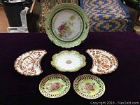 Full view of all six porcelain dishes arranged on dark cloth: one large handled plate, one 7-inch plate, two small floral pierced plates, and two red and white fan-shaped bone dishes.