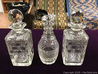 Lot of three vintage crystal decanters, two square and one rounded, all with stoppers, placed on dark surface showing overall set condition and shapes.