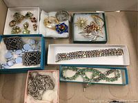 Image showing multiple jewelry pieces in boxes including bracelets, earrings, brooches, and shoe buckles on a beige surface.