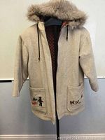 Full view of beige wool coat with fur-trimmed hood on black stand, showing front pockets and partially open to show quilted lining.