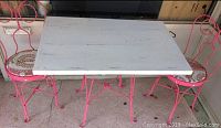 Vintage ice cream parlour table with white plank wooden top and pink wrought iron legs with two matching pink wrought iron chairs on a carpeted floor.