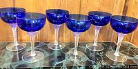 Six cobalt blue glass goblets arranged in a row on a marble or granite surface with wooden backdrop.