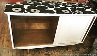 Full front view of credenza showing white frame, sliding door, open shelving space, and black and white floral patterned top.
