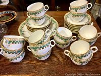 Porcelain cups stacked, displaying green and gold decorative pattern with white base. Variety of cup handle styles shown.
