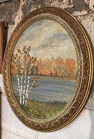 Oval oil painting showing a scenic landscape in autumn colors with birch trees, a lake, and cloudy sky in a decorative gold frame.