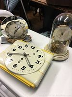 Three vintage clocks shown on a white table: white ceramic wall clock with yellow accents, silver and black desk clock, glass dome clock with brass mechanism