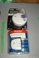 Photo of Kidde talking smoke and carbon monoxide alarm in original purple and black packaging and First Alert carbon monoxide alarm in blue and clear packaging