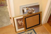 Group view of three framed prints leaning against a wall on wood floor, including a cat print, a print with children, and an empty gold ornate frame with backing.