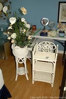 Wicker plant stand with faux flowers beside the white wicker shelving unit. Part of the room including floor and table with other items also visible.