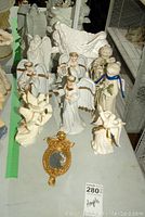 A group of angel figurines displayed on a table along with a small framed mirror and other figurines. The angels are mostly white with gold detailing on wings and robes.