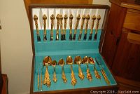 Open wooden case showing full set of gold-plated flatware arranged in teal fabric-lined slots.