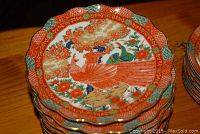 Close-up of stacked decorative plates with intricate bird and floral patterns, vibrant orange, green and blue colors, scalloped edges visible.