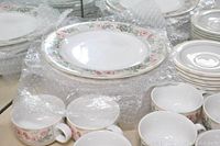 Photo showing stacked plates, cups, saucers and bowls with floral pattern on white ceramic dishes arranged on surface with bubble wrap nearby.