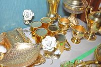 Photo showing gold tone coffee set including pot, cream, sugar, mugs, decorative white rose, lidded candy dish, large bowl, and tray on table.