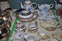Top-down view of multiple ceramic tea pots, creamers, sugar bowls, cups and saucers arranged on a table with floral and gold accents, including a heart-shaped sugar bowl with spoons.