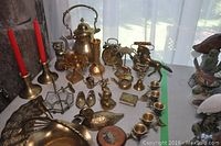 Table display of assorted brass decorative items including kettle, clock, candle holders, figurines, fan, and ashtrays, showing polished surfaces and various shapes in natural light.