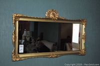 Full view of the rectangular framed mirror hanging on a blue wall, showing the gold ornate frame and its decorative accents.