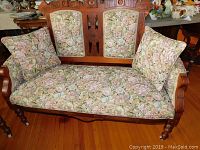 Close up of upholstered wooden settee showing floral pattern fabric cushions and carved wood frame details on backrest and arms.